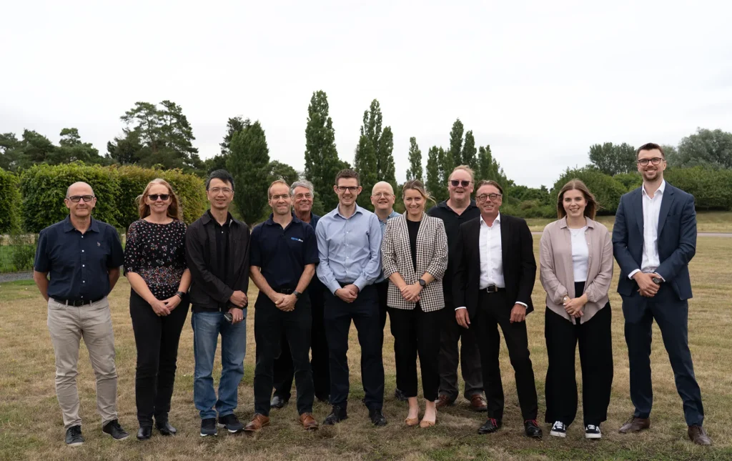 Group photo of twelve CML Micro colleagues smiling and standing in a line outdoors on grass, with trees in the background