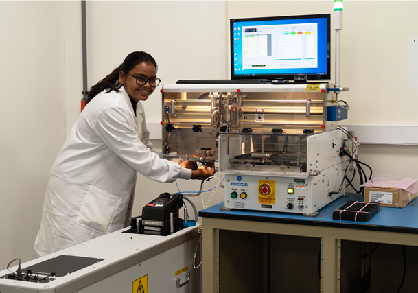 Woman in a lab coat operating a wire bonding machine in a laboratory, with a monitor displaying software readouts behind her